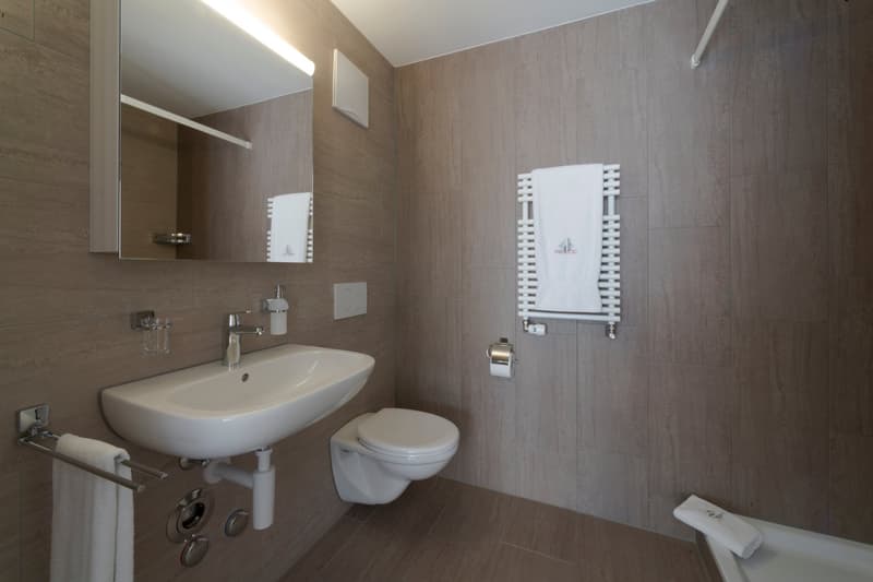 The image shows a modern bathroom with a white sink, toilet, and towel warmer. The walls are covered in a light-colored tile or panel material, creating a clean and minimalist aesthetic.