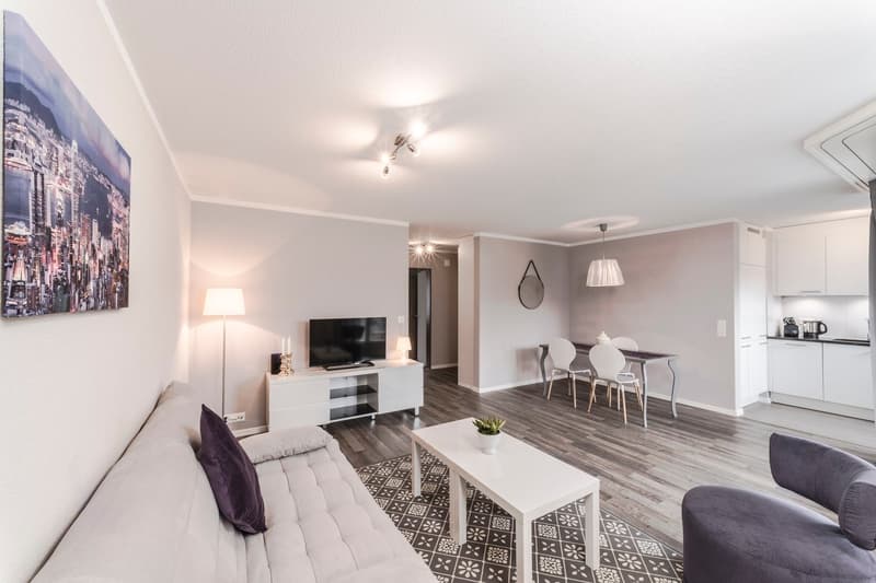 Spacious open-plan living room with hardwood flooring, white walls, large windows, a sofa, a TV stand, and a dining area with a table and chairs. The room also features a patterned area rug and a decorative mirror on the wall.