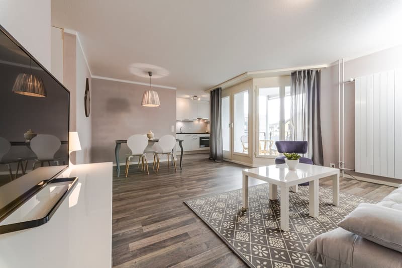 Open-concept living room and dining area with hardwood floors, gray walls, pendant lighting, white dining table and chairs, sofa, coffee table, and patterned area rug. Large windows provide natural light and access to a balcony or terrace.