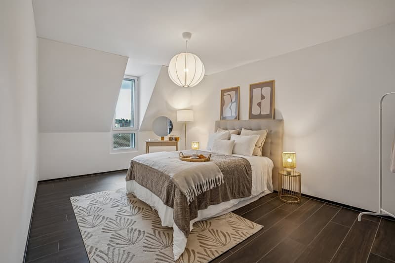 A spacious bedroom with white walls, a wooden floor, and a bed with a beige and brown bedspread. A hanging lamp with a white shade is fixed to the ceiling, while table lamps are placed on either side of the bed. A wooden nightstand with a lamp, a tray of
