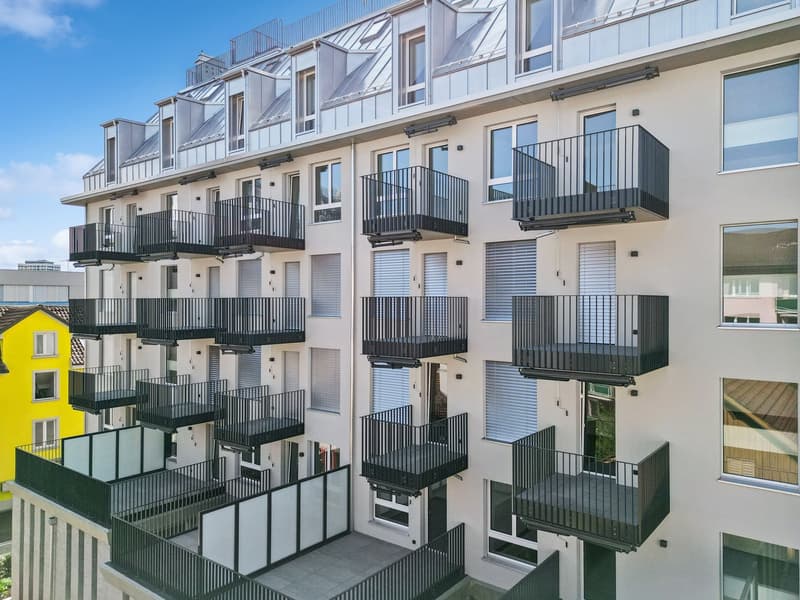 Modern white building with multiple balconies, large glass windows, and a yellow building next to it