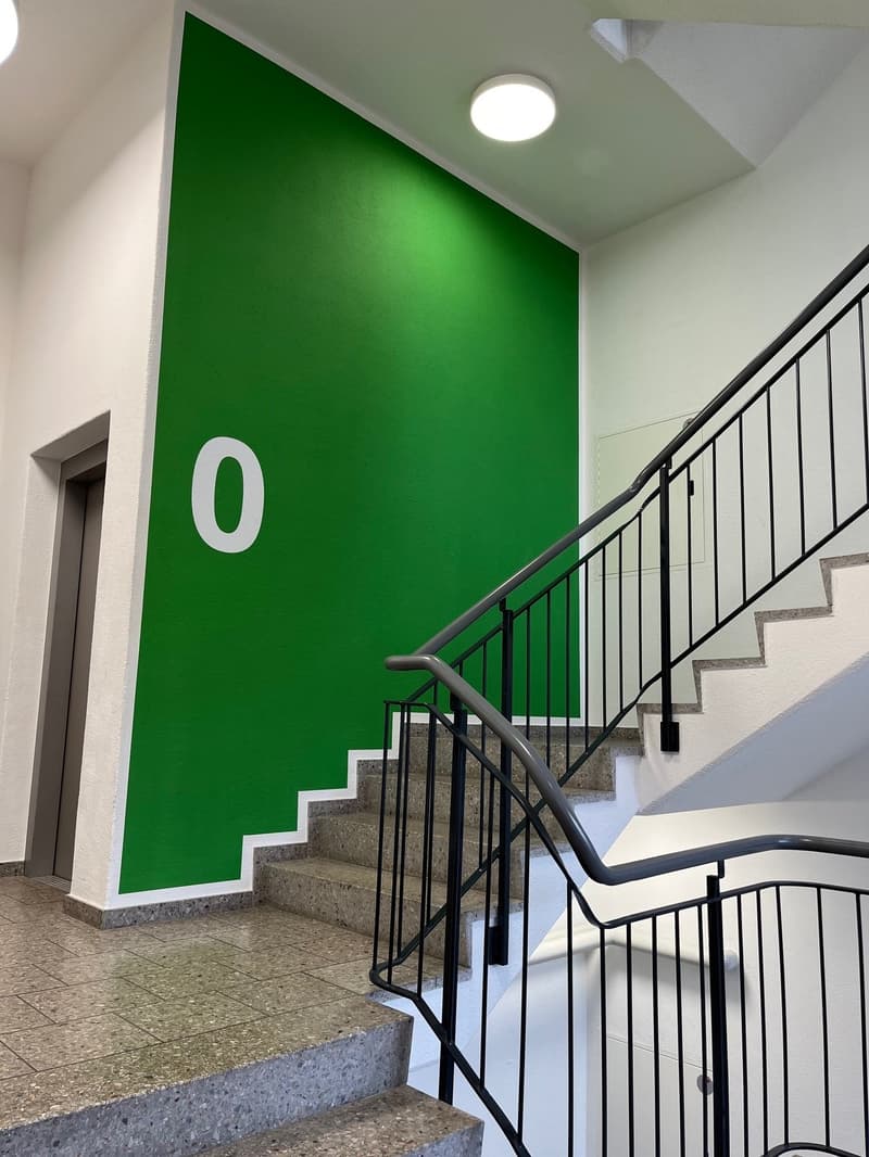 The image shows a staircase with a large green wall displaying the number 0. The stairs are made of concrete and have a metal railing. The lighting in the stairwell is provided by a ceiling-mounted light fixture.