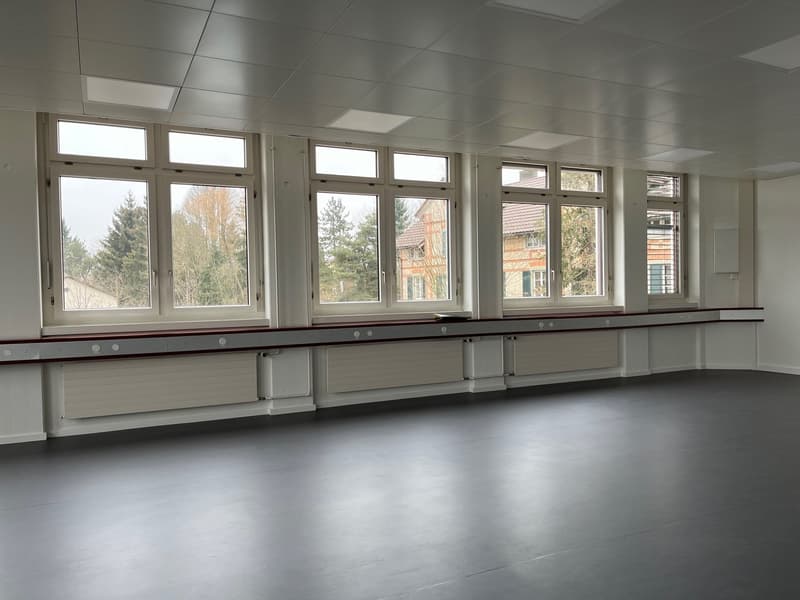 The image shows a large, open classroom with multiple windows along the walls, providing natural light. The room has a tiled floor and a suspended ceiling with recessed lighting. There is a long counter or bench running along one side of the room, likely
