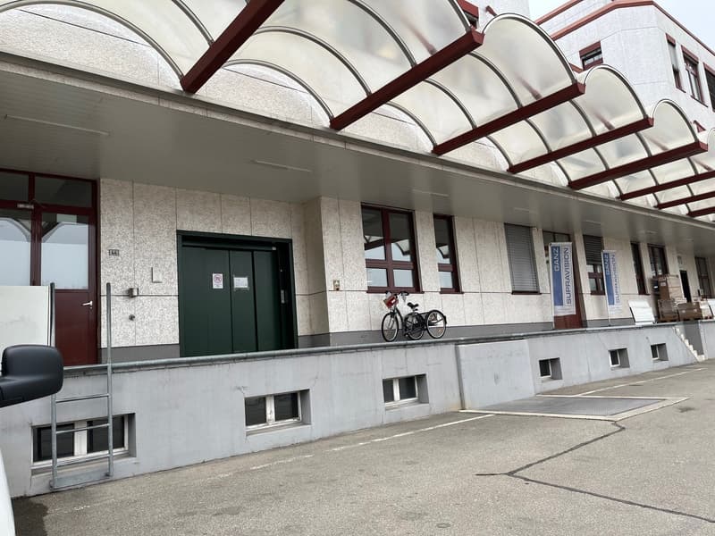 The image shows the exterior of a large, multi-story building with a distinctive arched glass roof structure. The building has a modern, minimalist design with a mix of concrete and glass materials. There is a bicycle parked outside the building's entranc