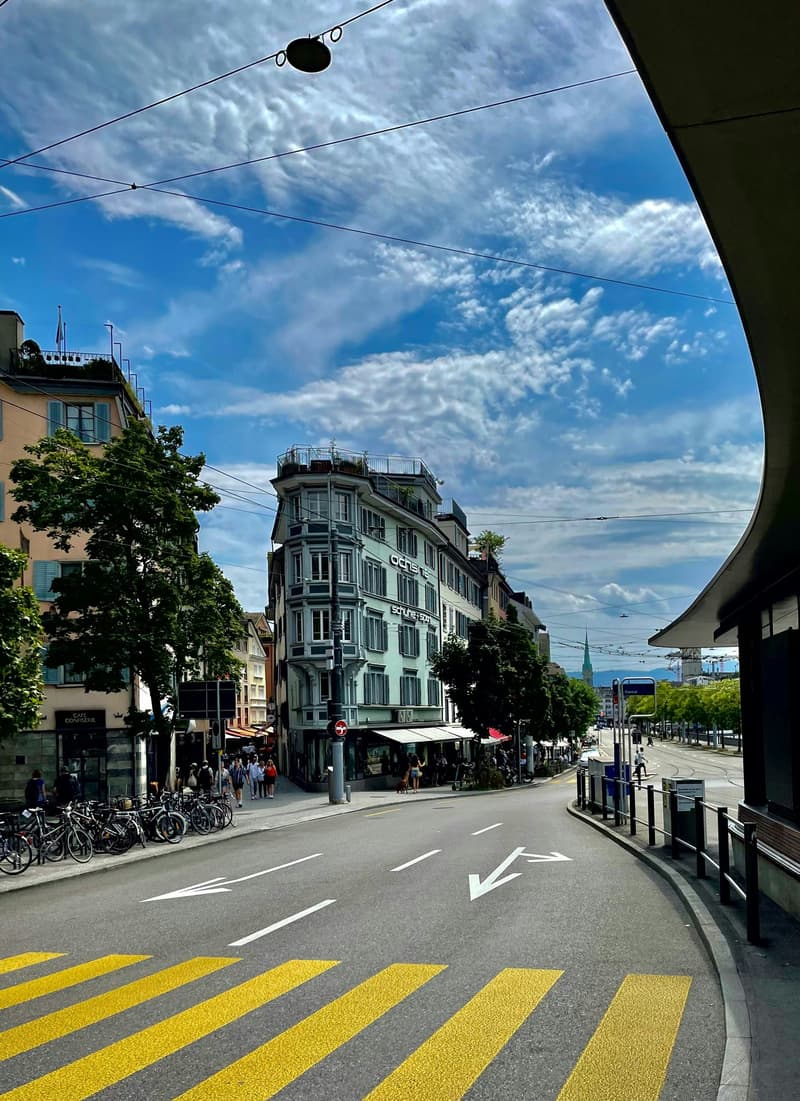 wide street, large modern building, lots of pedestrians, bike rental station, trees, clear blue sky