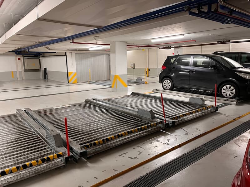 underground parking with two cars, concrete floors, metal barriers, ceiling beams, pipes, yellow and black striped poles