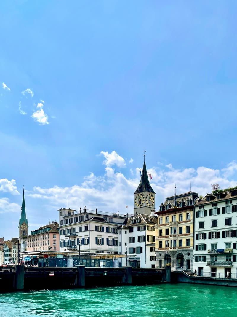 Cityscape view with buildings along the river, one with a clock tower, under a partly cloudy sky.