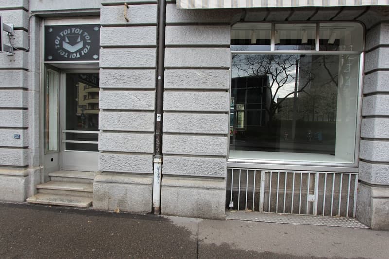 Gray stone building with two entrances, one with stairs and one with a glass door, large glass windows, metal grates