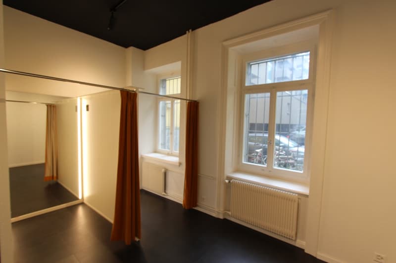 Empty room with white walls, black floor, large mirror, two windows with orange curtains, radiators