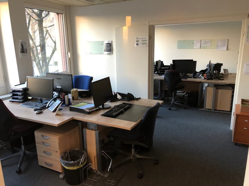The image shows an office space with multiple desks, computers, and office equipment. The room has large windows providing natural light, and the walls are painted a light color. There are various office supplies and documents visible on the desks.