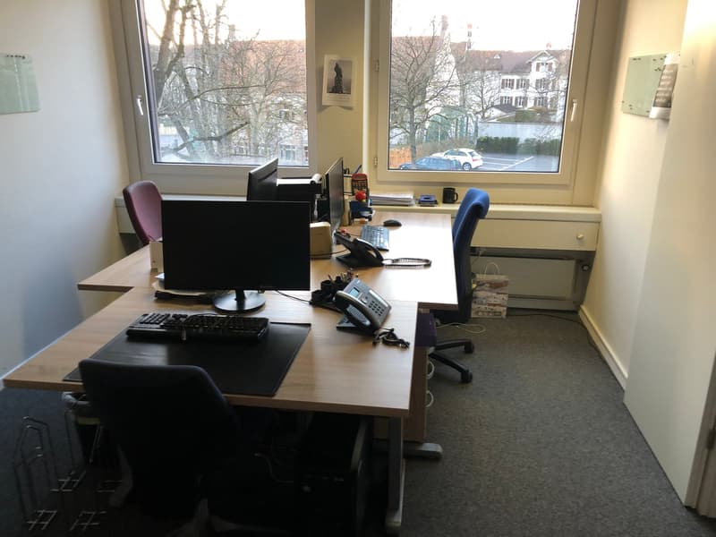 The image shows an office workspace with a desk, computer monitor, keyboard, phone, and other office equipment. The room has large windows overlooking a street outside with trees and buildings visible. The walls are light-colored and the floor appears to