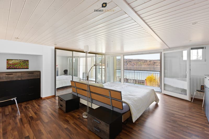 Jeden Morgen erwachen und den Blick über den glitzernden See schweifen lassen - ein Genuss, der das Herz berührt. / a modern master bedroom with a large bed, hardwood floors, and large windows tha