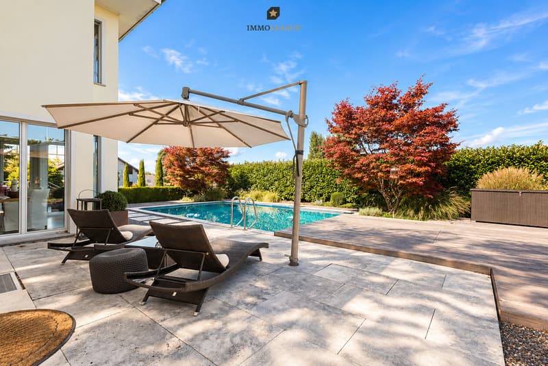 der Garten eines modernen Hauses mit einem großen Swimmingpool und einer Terrasse mit Stühlen und einem Sonnenschirm / the backyard of a modern home with a large swimming pool and patio with chairs