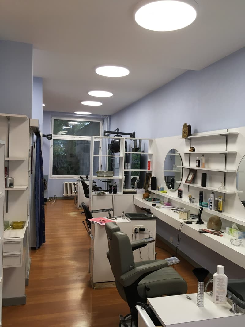 The image shows a beauty salon or hair salon with various equipment and supplies. The room has a modern and organized layout, with shelves and storage units lining the walls. There are several salon chairs and a dishwasher visible, indicating this is a pr