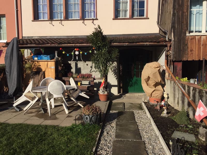 Apartment building exterior with a covered patio, a table and chairs, a wooden cabinet, a green door, a plant in a pot, and decorative ornaments.