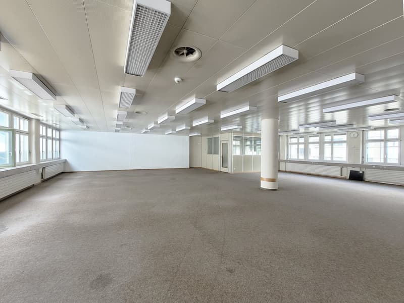 Large open office space with gray carpeted floor, white walls, and multiple ceiling lights. The room has several windows along the walls and a few columns in the center.