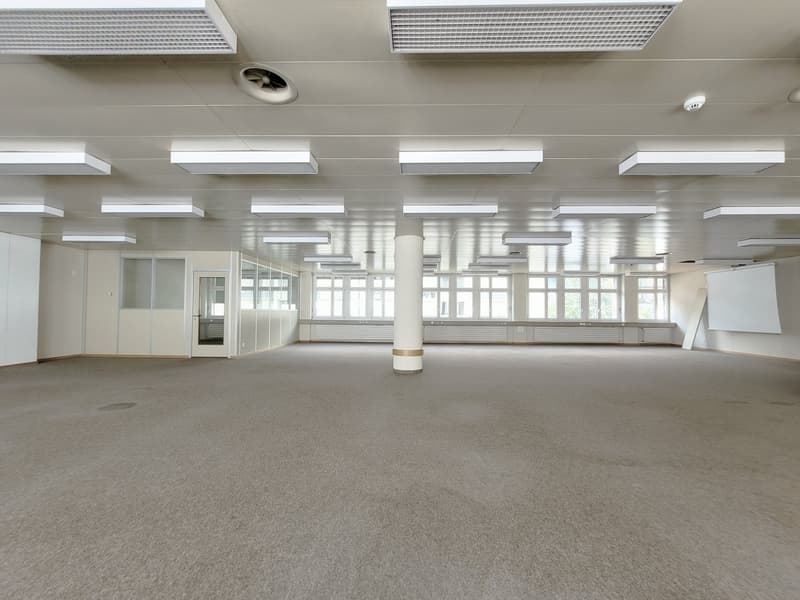 Large open office space with high ceilings, fluorescent lighting, and windows along the walls. The room has a carpeted floor and appears to be empty, ready for use.