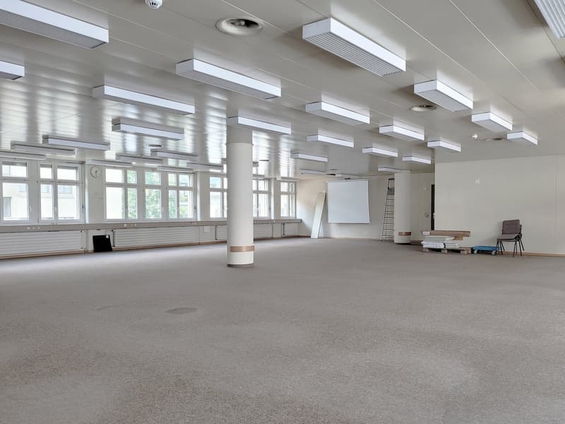 Large open room with high ceilings, multiple windows, and a whiteboard. The room has a gray carpeted floor and is equipped with various lighting fixtures and furniture, including chairs and a ladder.