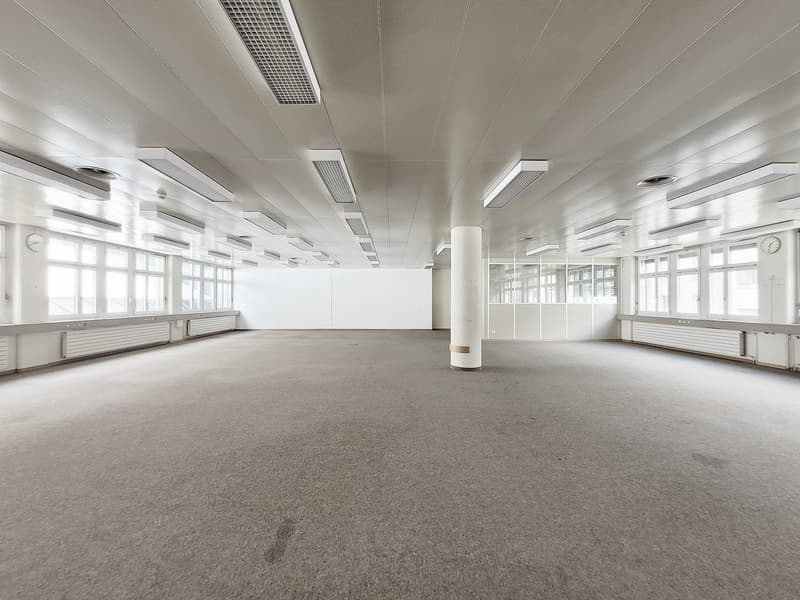 Large open office space with high ceilings, white walls, and gray carpeted flooring. The room has multiple windows along the walls, providing natural light. There are several ceiling-mounted lights and air conditioning units installed. The space appears t