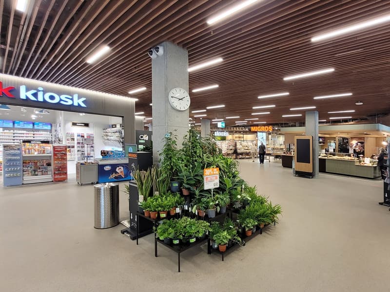 The image shows the interior of a supermarket with a Kiosk counter and a Migros section. The ceiling has a wooden slat design with recessed lighting. There is a large clock mounted on a column in the center of the space. The floor is tiled and there are v
