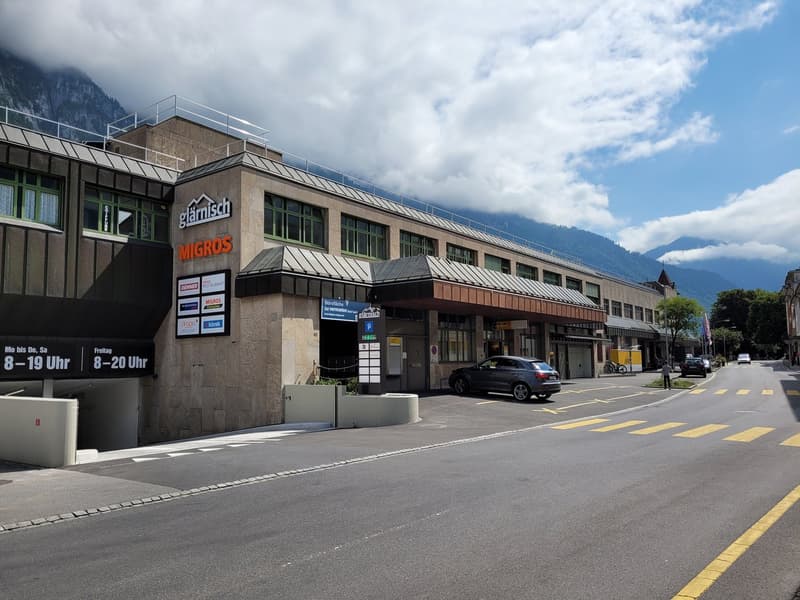 A large commercial building with a Migros supermarket and other stores. The building has a modern, rectangular design with large windows and a metal roof. There is a parking area in front of the building and the surrounding area appears to be a city or to