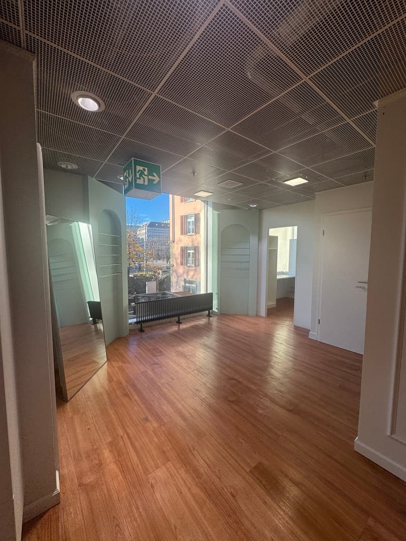 An empty room with a wooden floor, two mirrors on the left side, and an exit sign on the ceiling. It has windows overlooking a balcony with a railing and buildings in the distance.