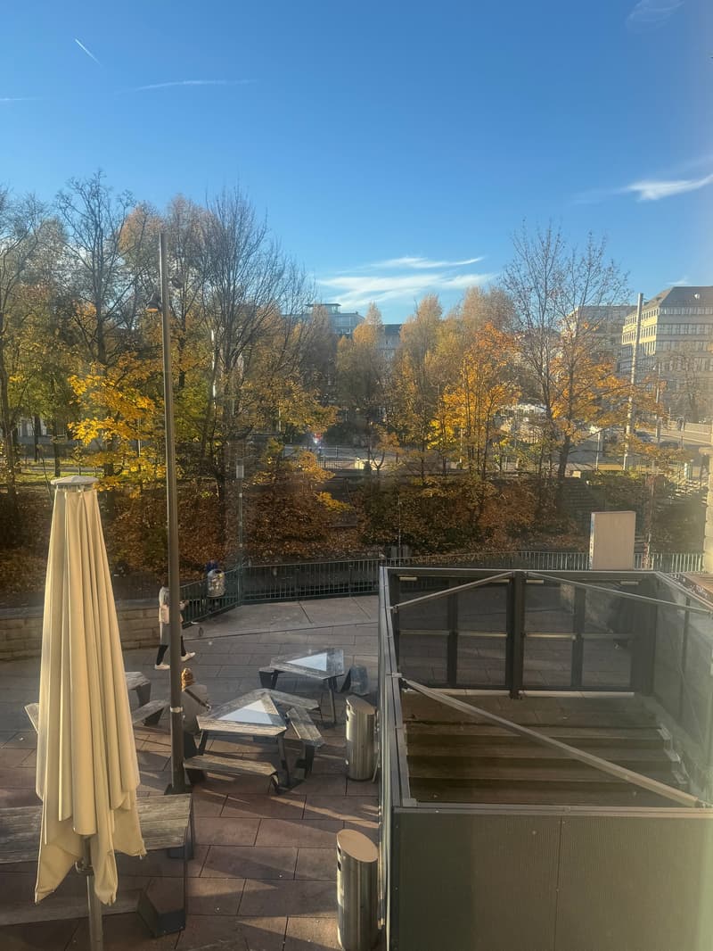 outdoor seating area with metal tables, benches, and umbrellas, fenced balcony with railings, open view of a park with trees, people walking
