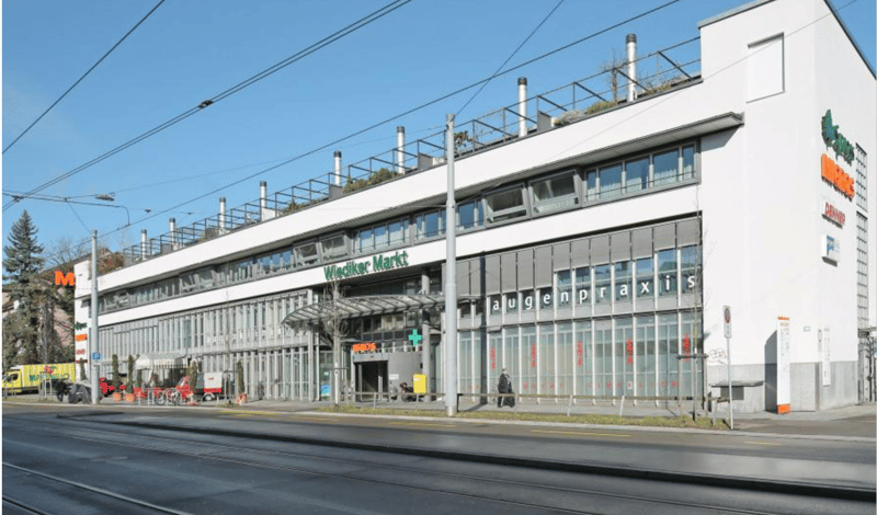 Modern white building with glass windows, multiple floors, green signboard, entrance with canopy, streetlights, and a tram line above