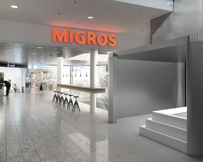 Entrance of Migros store, modern design, tiled floor, bar counter, stools, signage, pharmacy section, ATM machine, person standing
