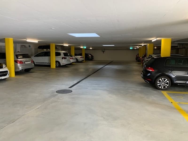 The image shows an indoor parking garage with multiple levels. The floor is made of concrete and there are yellow support columns throughout the space. Several cars are parked in the garage, including SUVs and sedans.