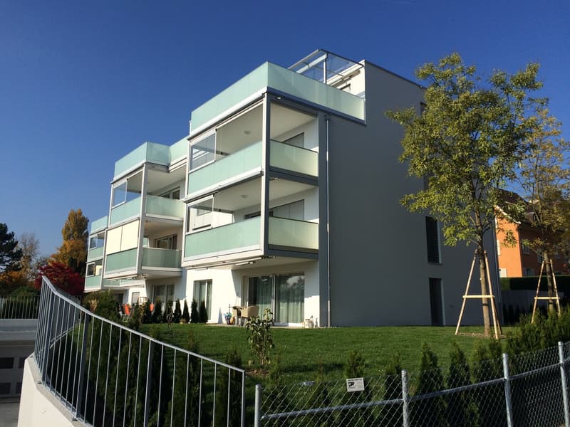 Modern multi-story apartment building with balconies, glass windows, and a landscaped yard with trees and shrubs.