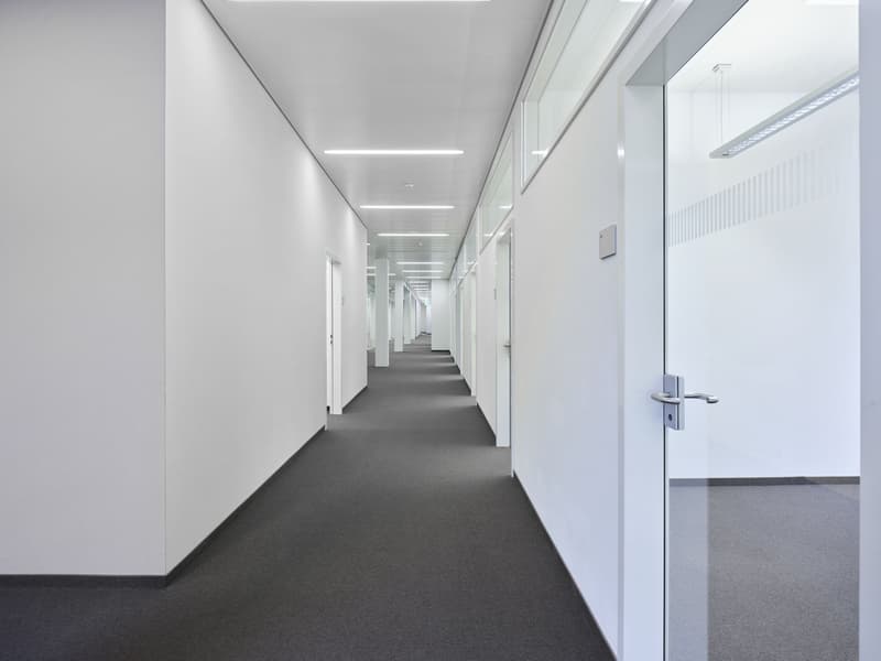 The image shows a long, well-lit office corridor with white walls and a gray carpet floor. The corridor has multiple doors lining the walls, indicating individual office spaces. The lighting fixtures on the ceiling provide a bright and uniform illuminatio