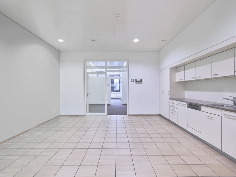 Large open office space with white walls, tile flooring, and a kitchen area with cabinets and appliances. The space has recessed lighting and a glass door leading to another room.