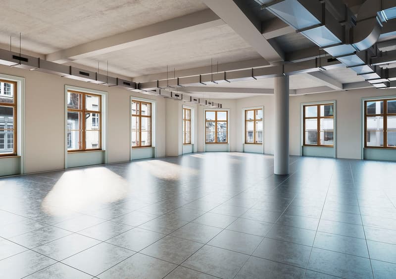 Large open-plan office space with high ceilings, large windows, and a modern, minimalist design. The room has a tiled floor and is equipped with hanging light fixtures. The walls are painted in a neutral color scheme, creating a bright and airy atmosphere