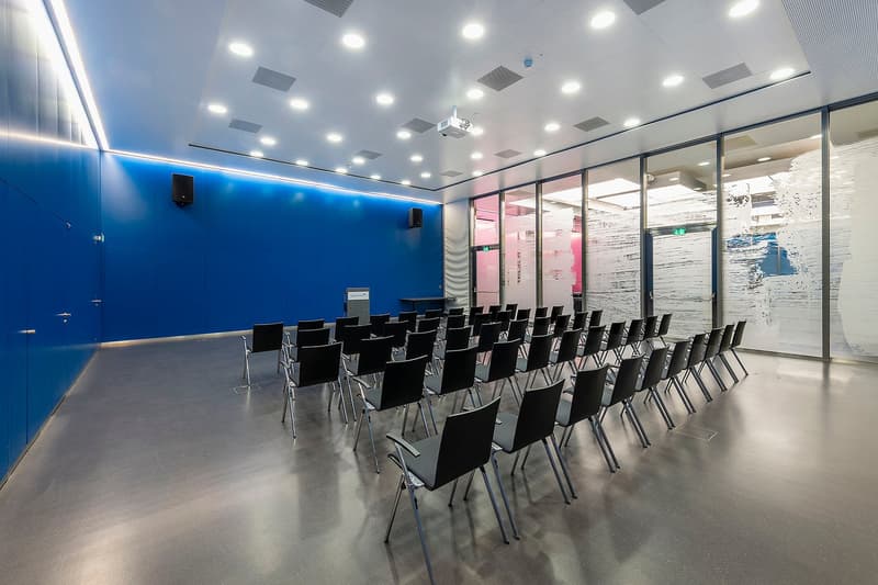 Large conference room with blue walls, rows of black chairs facing a projection screen, and recessed lighting in the ceiling.