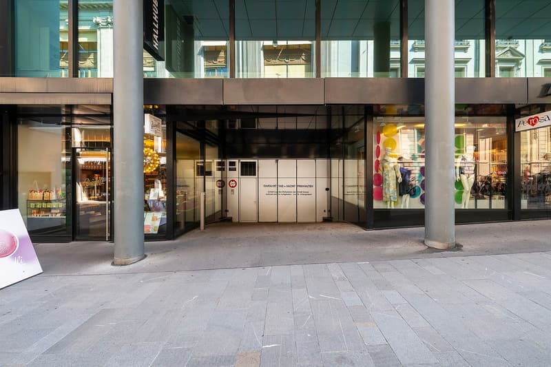 The image shows the exterior of a commercial building with large glass windows and doors. There are several retail stores and businesses visible, including a Pettosa store. The building has a modern, industrial-style architecture with exposed beams and co