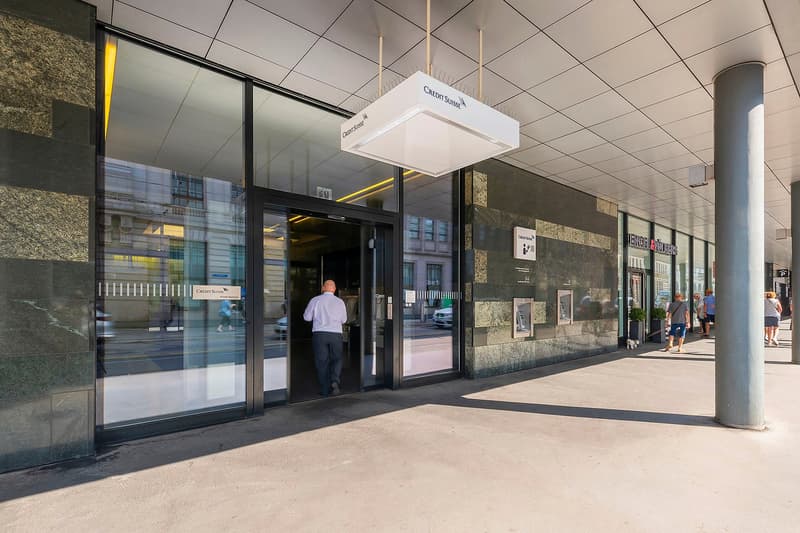 Exterior of a commercial building with a Credit Suisse sign, glass entrance doors, and a person entering the building. The building has a stone facade and there are other shops visible along the street.