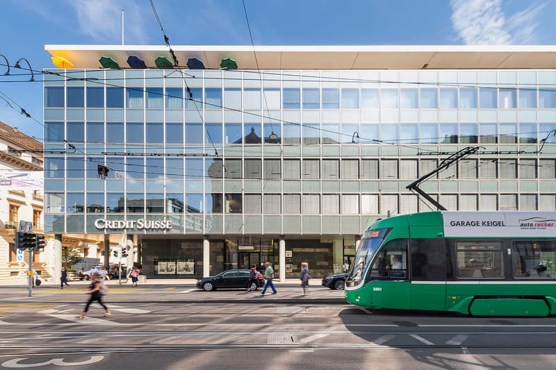 A multi-story commercial building with a Credit Suisse bank branch and a Garage Keigel auto repair shop on the ground floor. The building has a glass facade with a grid-like pattern. There are people walking on the sidewalk in front of the building, and a