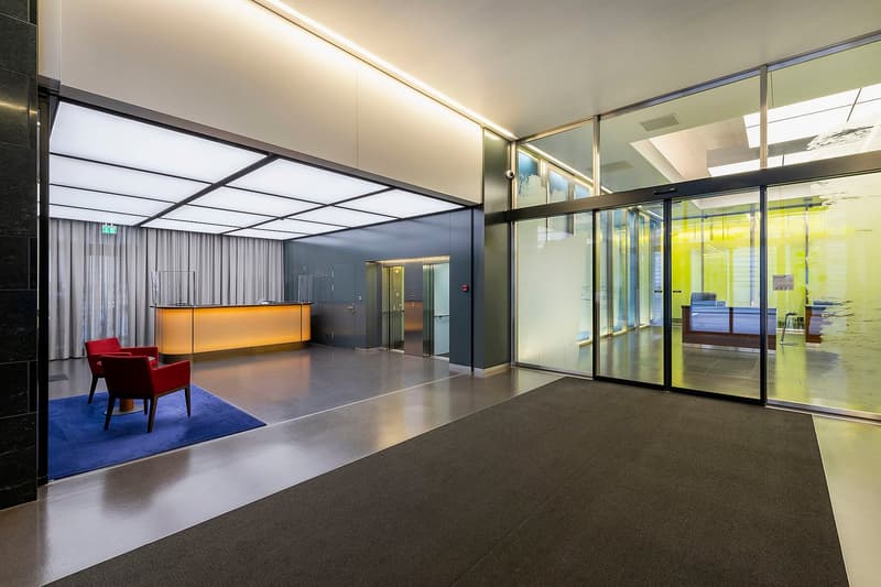 The image shows a modern and stylish office lobby. The space features a large reception desk with a golden-colored front, glass walls and doors, and a blue rug on the floor. The lighting is bright and the overall design has a sleek, contemporary feel.