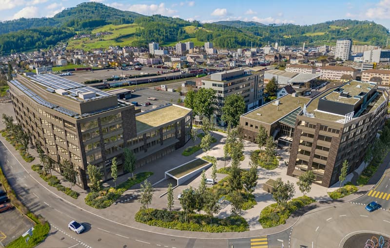 Modern office building with solar panels on the roof, surrounded by greenery and a parking lot.