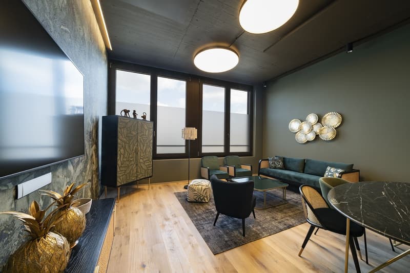 Open living area with large windows, wooden floor, dark green walls, modern furnishings, a green sofa, a round coffee table, armchairs, a floor lamp, decorative wall hangings, a large TV, two golden pineapple statues, and decorative pillows on the sofa.