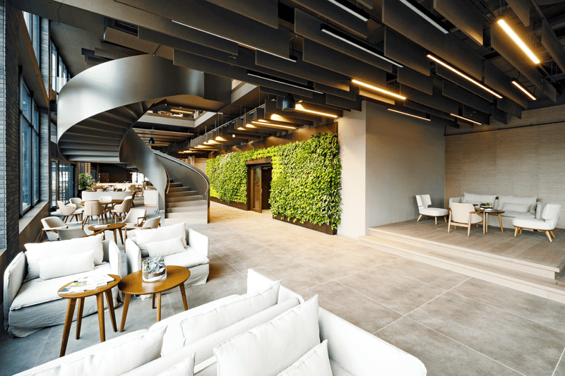 A contemporary indoor space with white furniture, wooden tables, large windows, a spiral staircase, a green wall, modern lighting, and a minimalistic aesthetic.