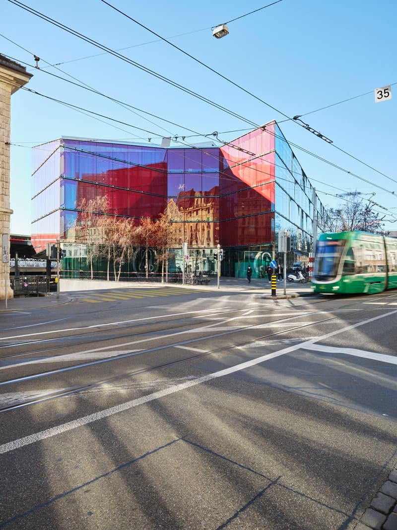 Modern building with glass facade, red and blue colors, located on a busy street with tram tracks and a green tram passing by.
