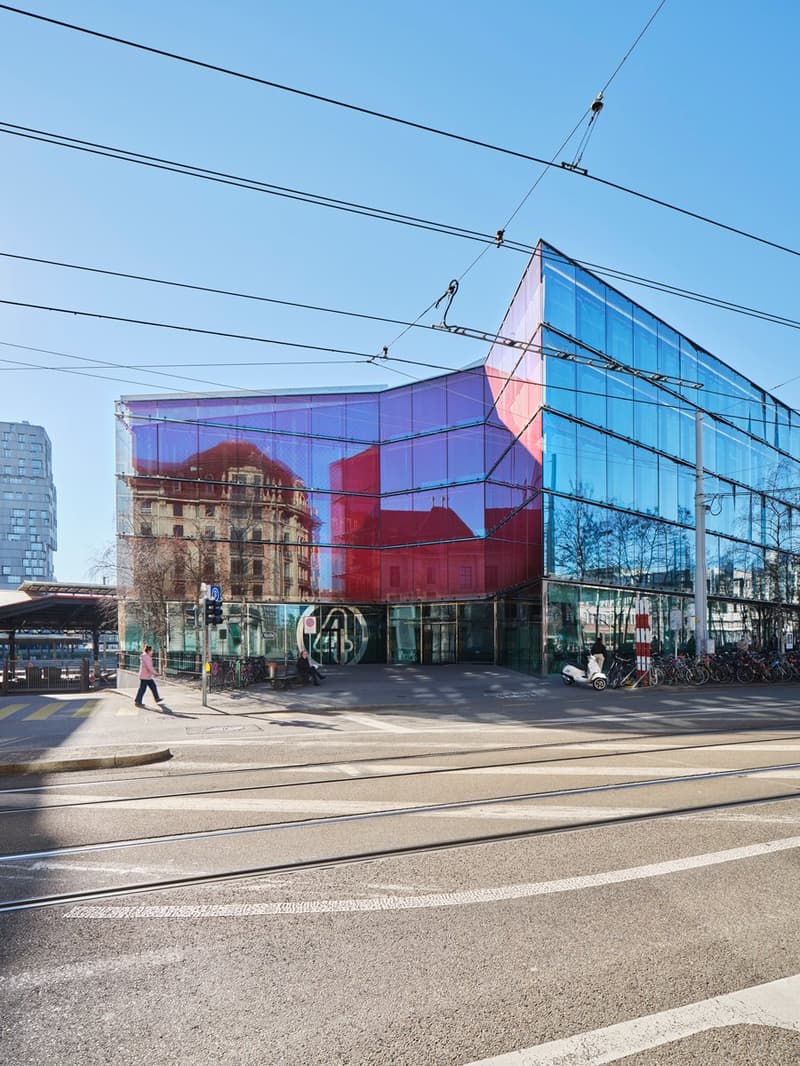 Modern building with glass facade, red and blue reflections, multiple windows, entrance with glass doors, bicycles parked in front, street with tram tracks, pedestrian crossing, clear sky
