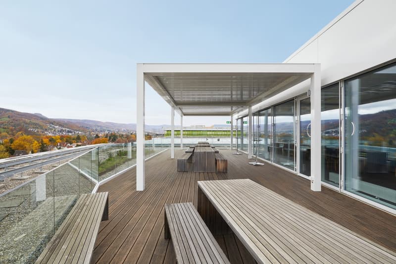 Wooden deck with benches and tables, glass railings, covered area with metal roof, scenic view of mountains and town