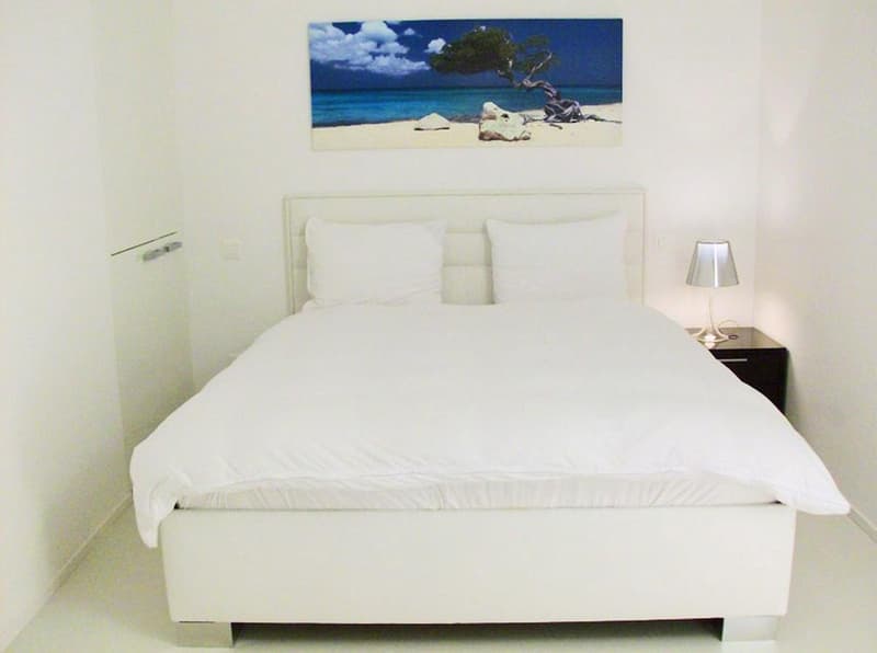 A modern white bedroom with minimalist design, featuring a large white bed, matching white pillows, a beach-themed painting, and a bedside table with a lamp.