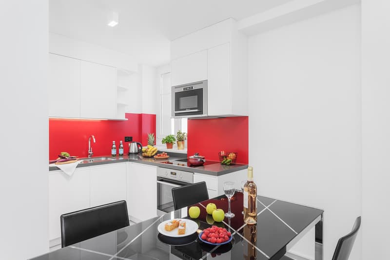 Modern white kitchen with red accent walls, built-in microwave, under-counter dishwasher, large dining table, high-gloss finish, stainless steel appliances, glass tabletop, wine bottle, glasses, fruit bowl
