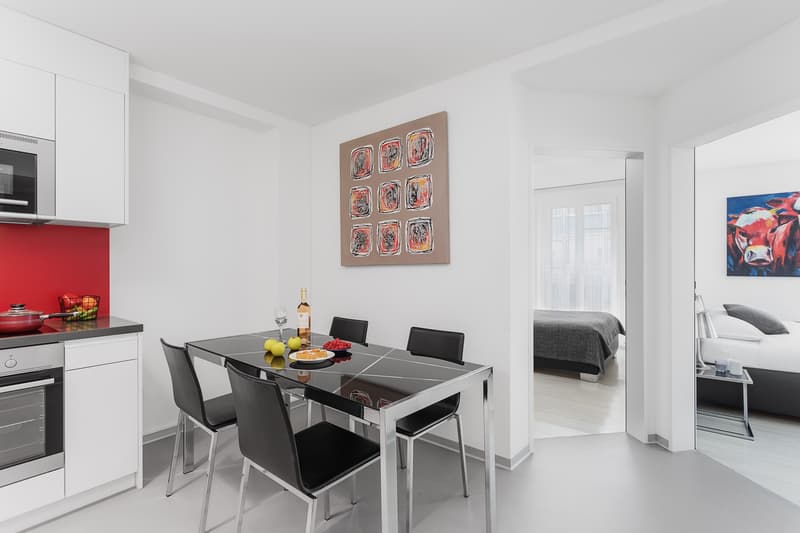 Modern dining area with glass table and black chairs, white kitchen cabinets, red kitchen wall, oven, microwave, dining table, wine bottle, glasses, and decorative items. There is a bedroom with a bed, pillows, and a painting in the distance.