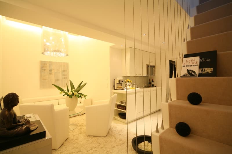 Minimalist living room with white furniture, beige carpet, white couch, glass table, a small flower vase, a white table, a white chair, a statue of Buddha, and a staircase leading to the next floor.