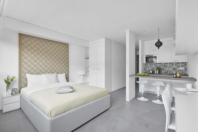 white interior, king size bed, 2 night tables, small kitchen, breakfast bar, 3 bar stools, round lamp, small plants, countertop vases